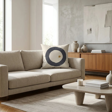 Modern cream cushion with a navy blue Enso circle design on a beige sofa in a minimalist living room setting with wooden furniture.