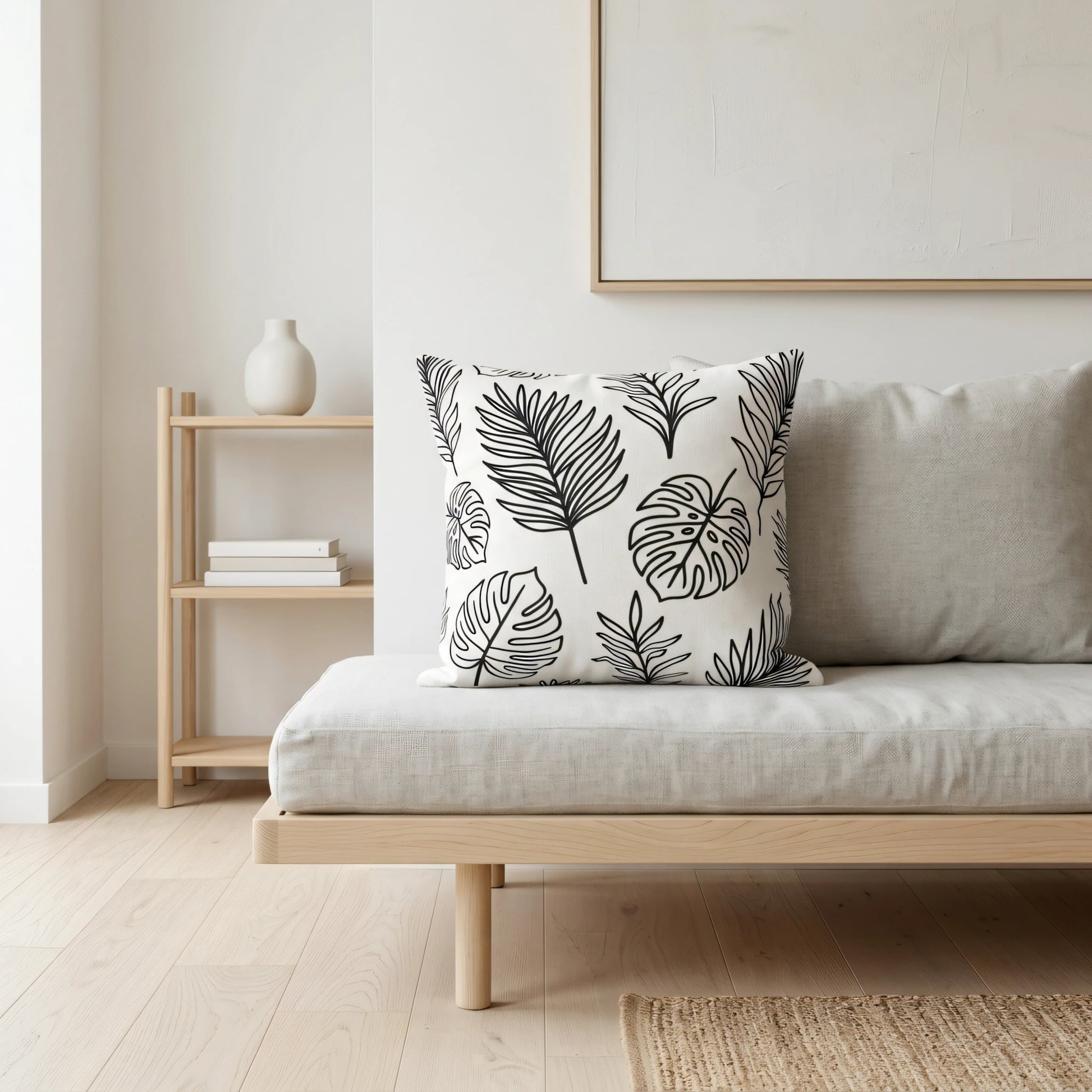 White decorative cushion with black botanical leaf patterns on a grey sofa in a minimalist living room with light wood furniture and neutral decor.