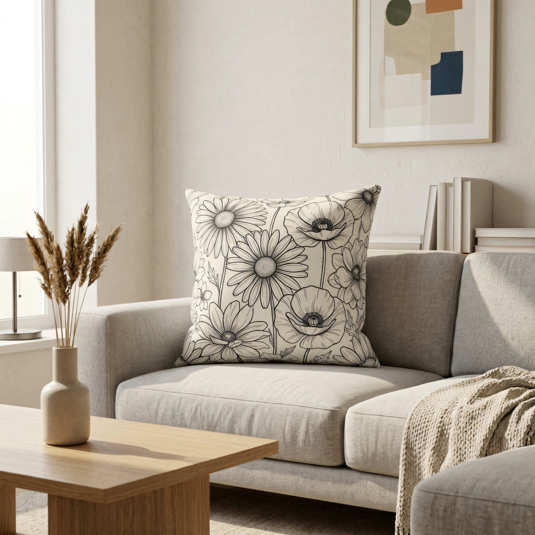 Modern floral throw pillow with black line art daisies on a cream background, placed on a grey sofa in a minimalist living room interior.