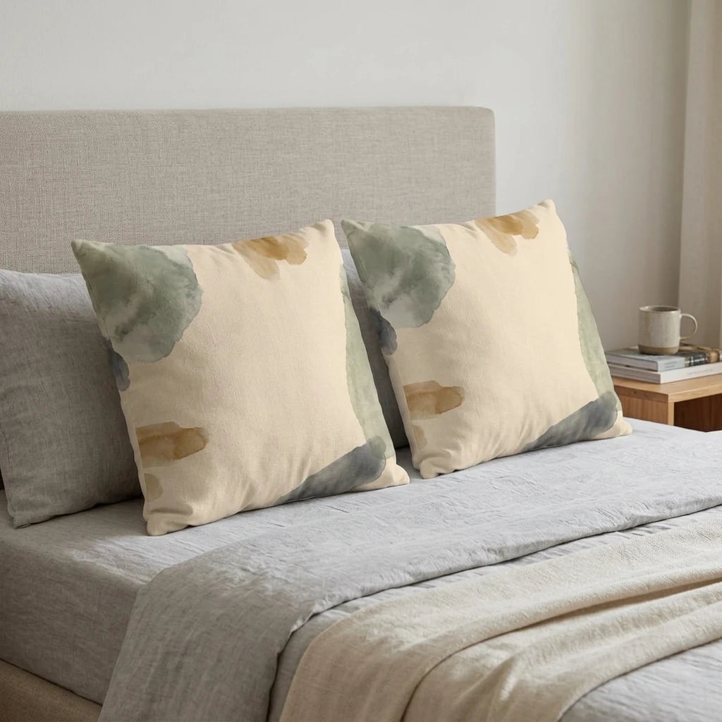 Two watercolor throw pillows with green and gold abstract designs on a neutral bed. Minimalist lifestyle bedroom decor with modern cushions.
