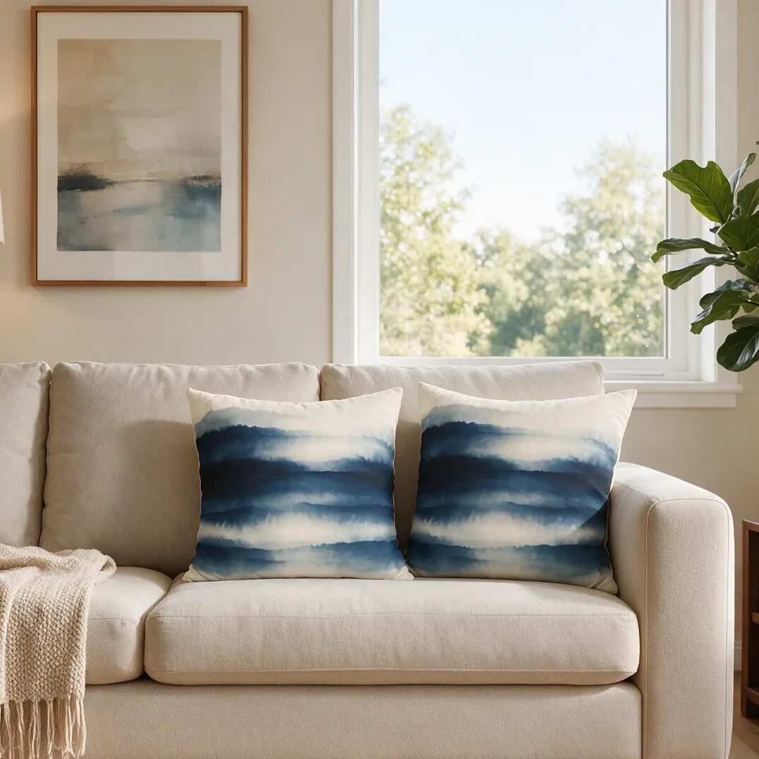 Pair of decorative cushions with blue watercolor stripes on a beige sofa in a bright living room setting with a framed art piece.