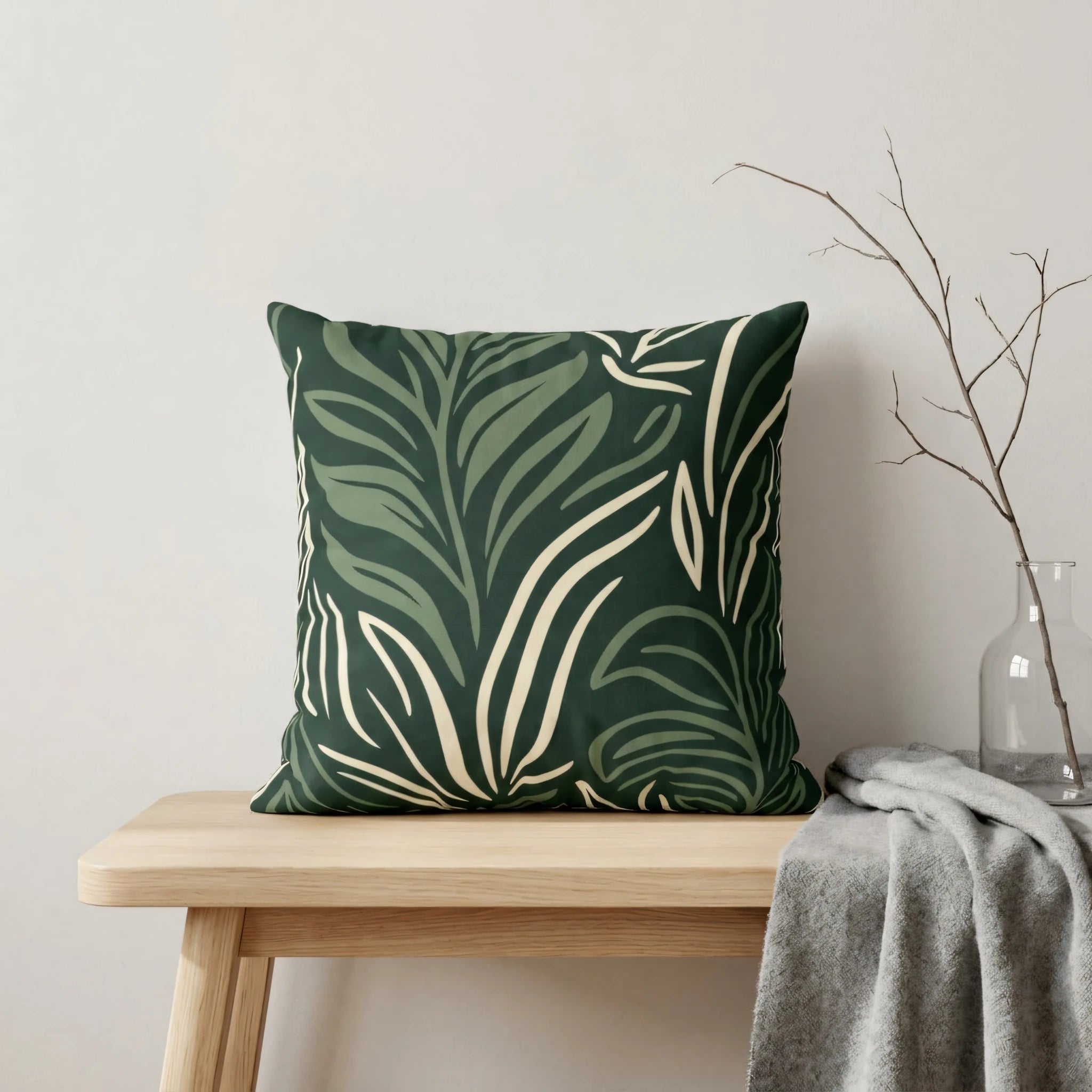 Decorative green leaf print pillow on a wooden bench in a minimalist interior with a gray throw and dried branch in a glass vase.