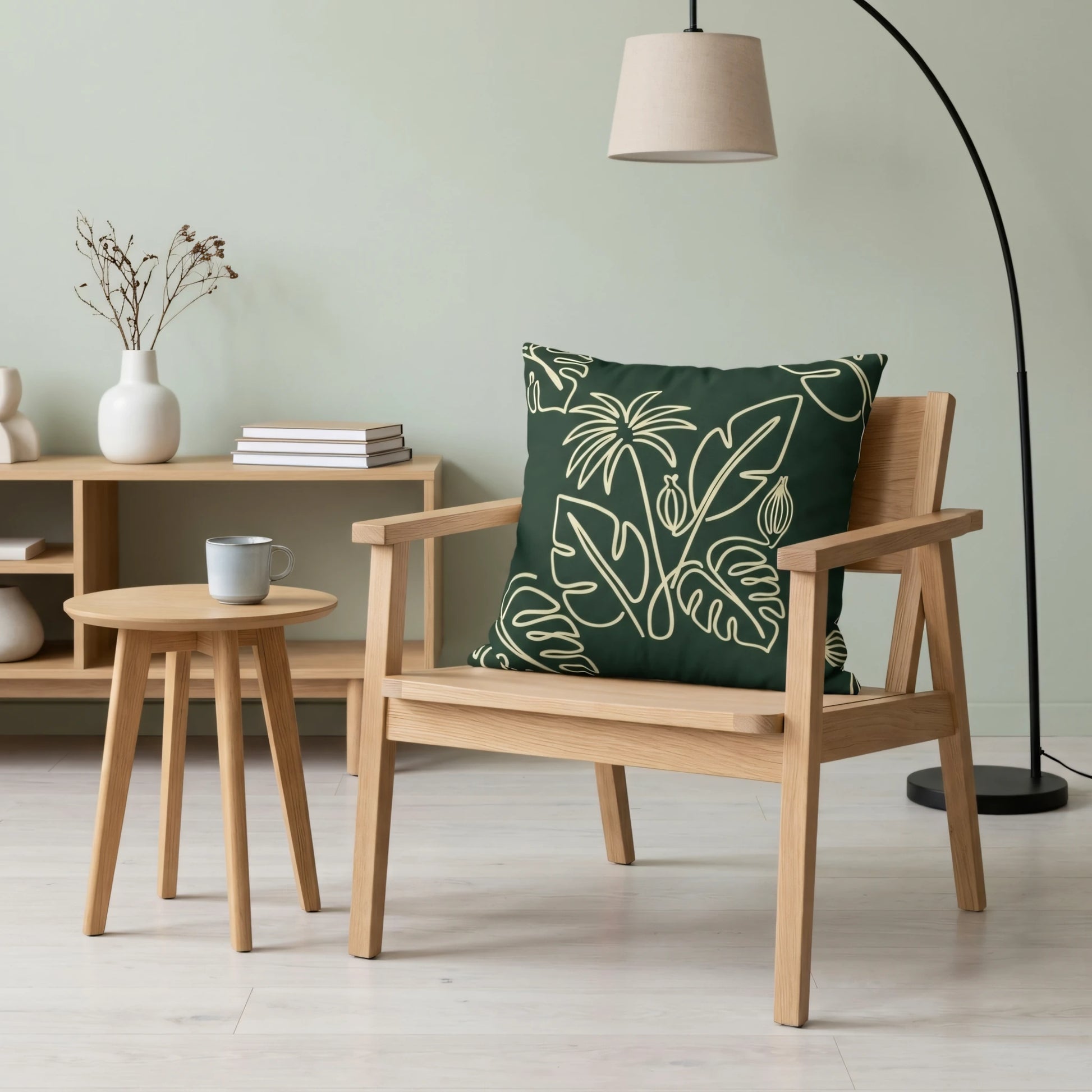 Dark green botanical throw pillow on a wooden armchair in a minimalist room with a side table, floor lamp, and sage green wall.