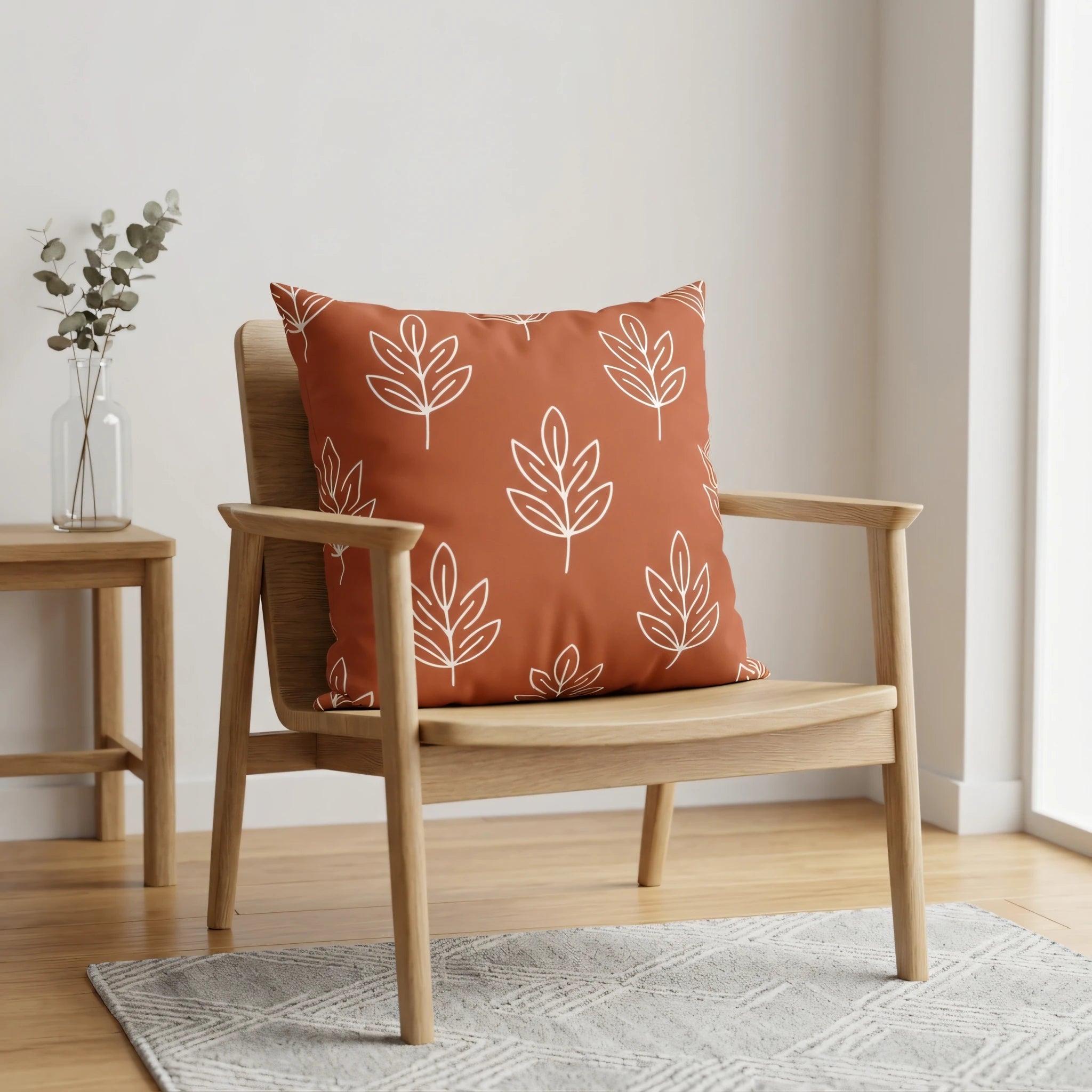 Terracotta throw pillow with white line-art leaf patterns on a wooden armchair in a bright minimalist room with a side table and rug.