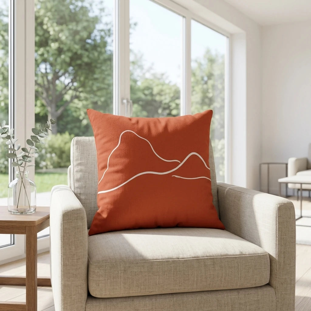 Burnt orange throw pillow with white mountain line art on a beige armchair in a bright sunlit room with large windows and minimalist decor.