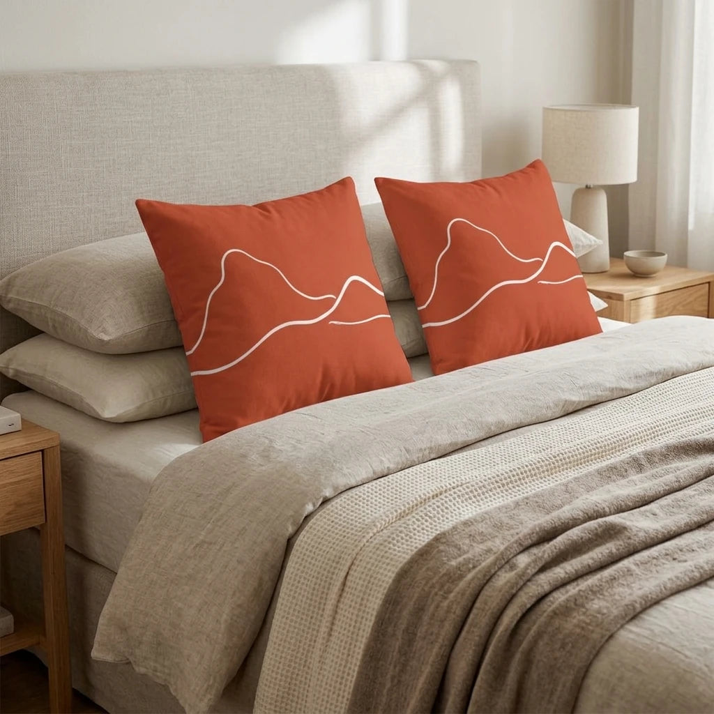 Two terracotta cushions with white minimalist mountain line art on a cozy bed with beige linens in a neutral bedroom.