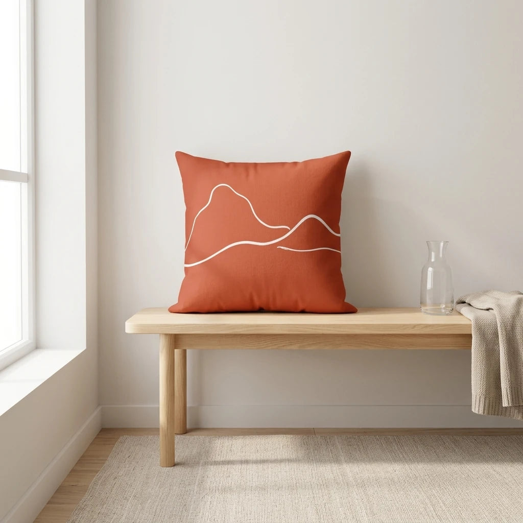 Terracotta throw pillow with white mountain line art on a wooden bench in a minimalist room with natural light and a cozy knit blanket.