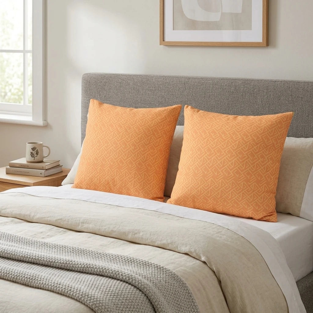 Two orange cushions with a geometric labyrinth pattern on a bed with a grey headboard and neutral bedding in a cozy bedroom setting.