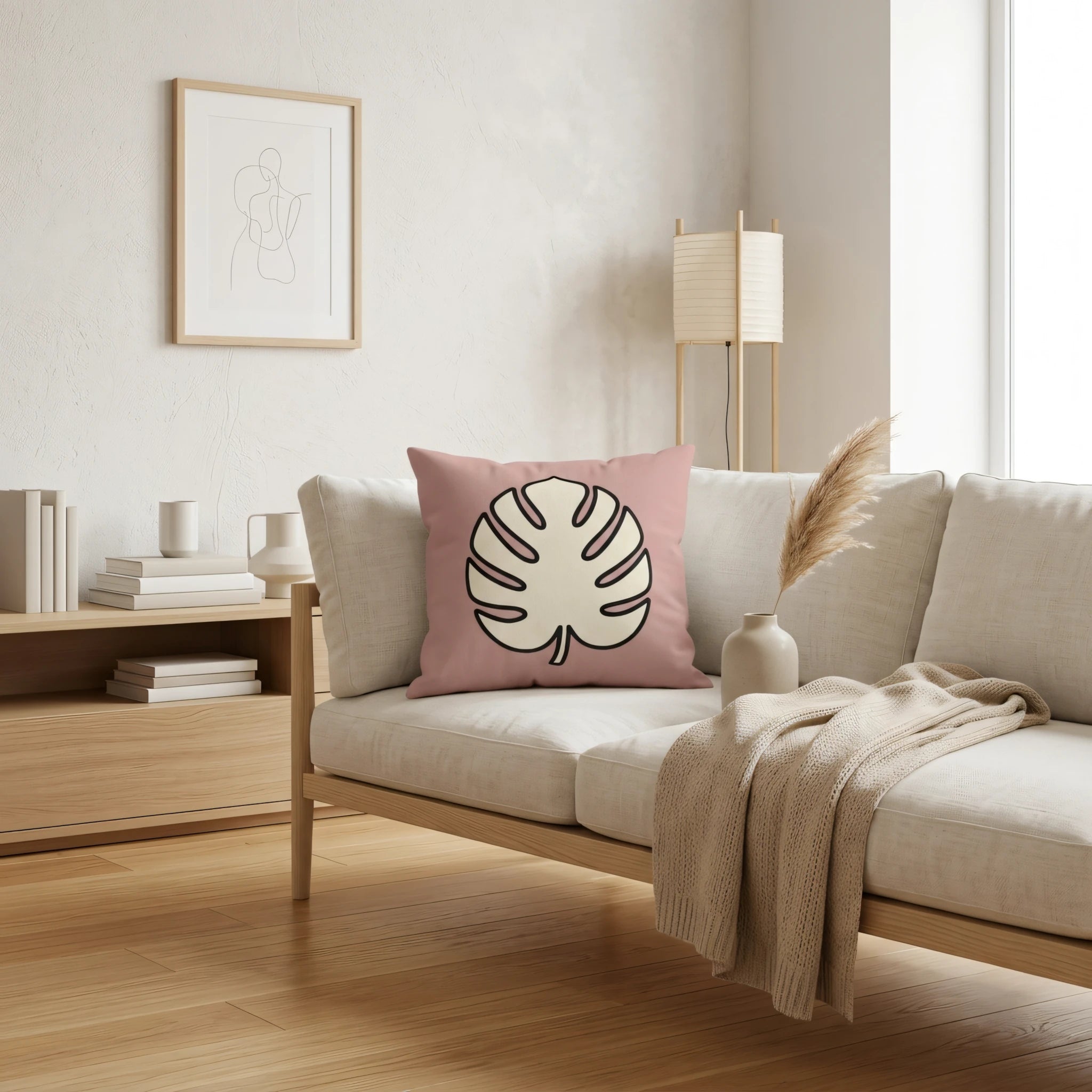 Dusty pink cushion featuring a monstera leaf design on a beige sofa in a minimalist interior with a wooden floor and simple wall art.