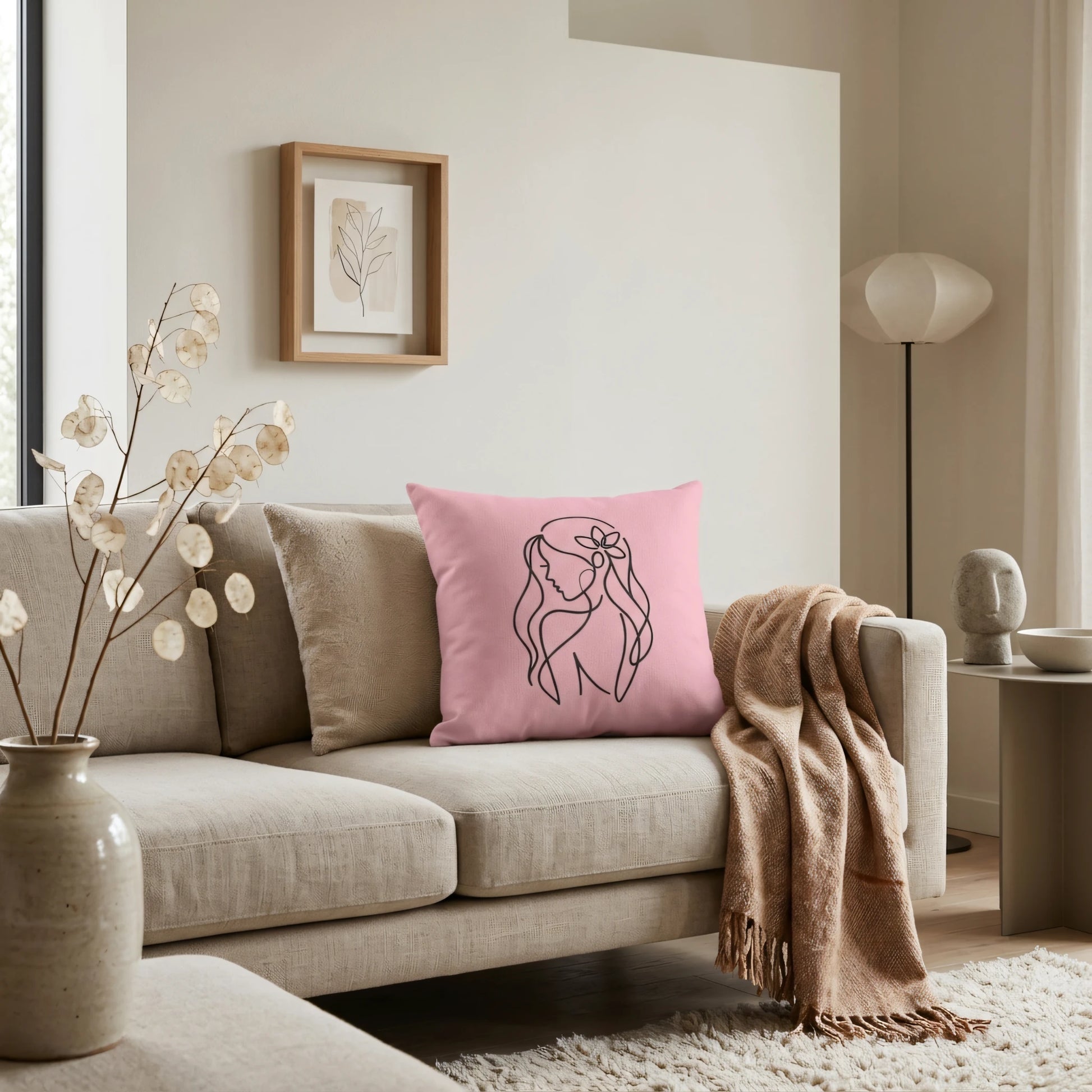 Pink throw cushion with black line art of a woman on a beige sofa in a minimalist living room interior with neutral decor accents.