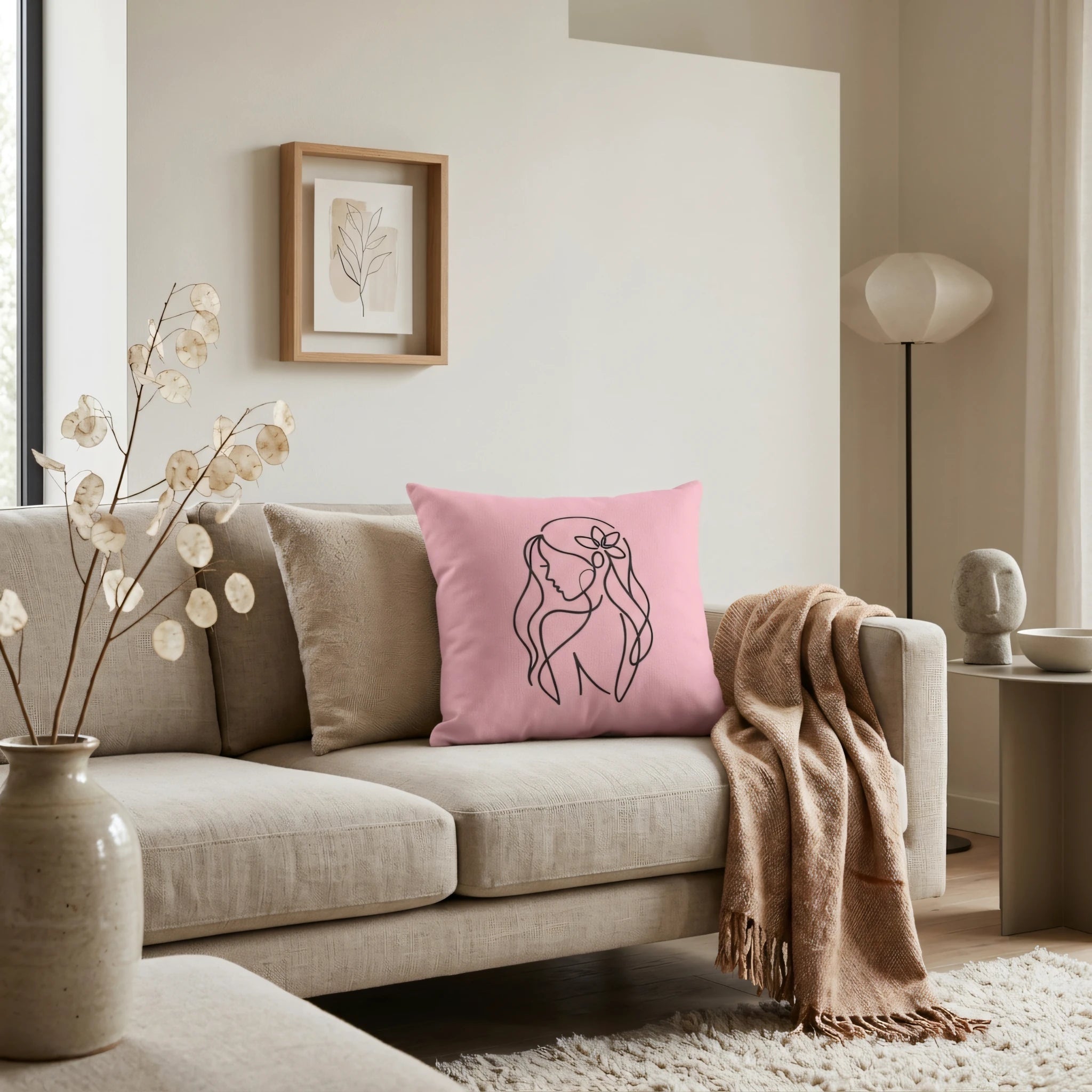 Pink throw cushion with black line art of a woman on a beige sofa in a minimalist living room interior with neutral decor accents.