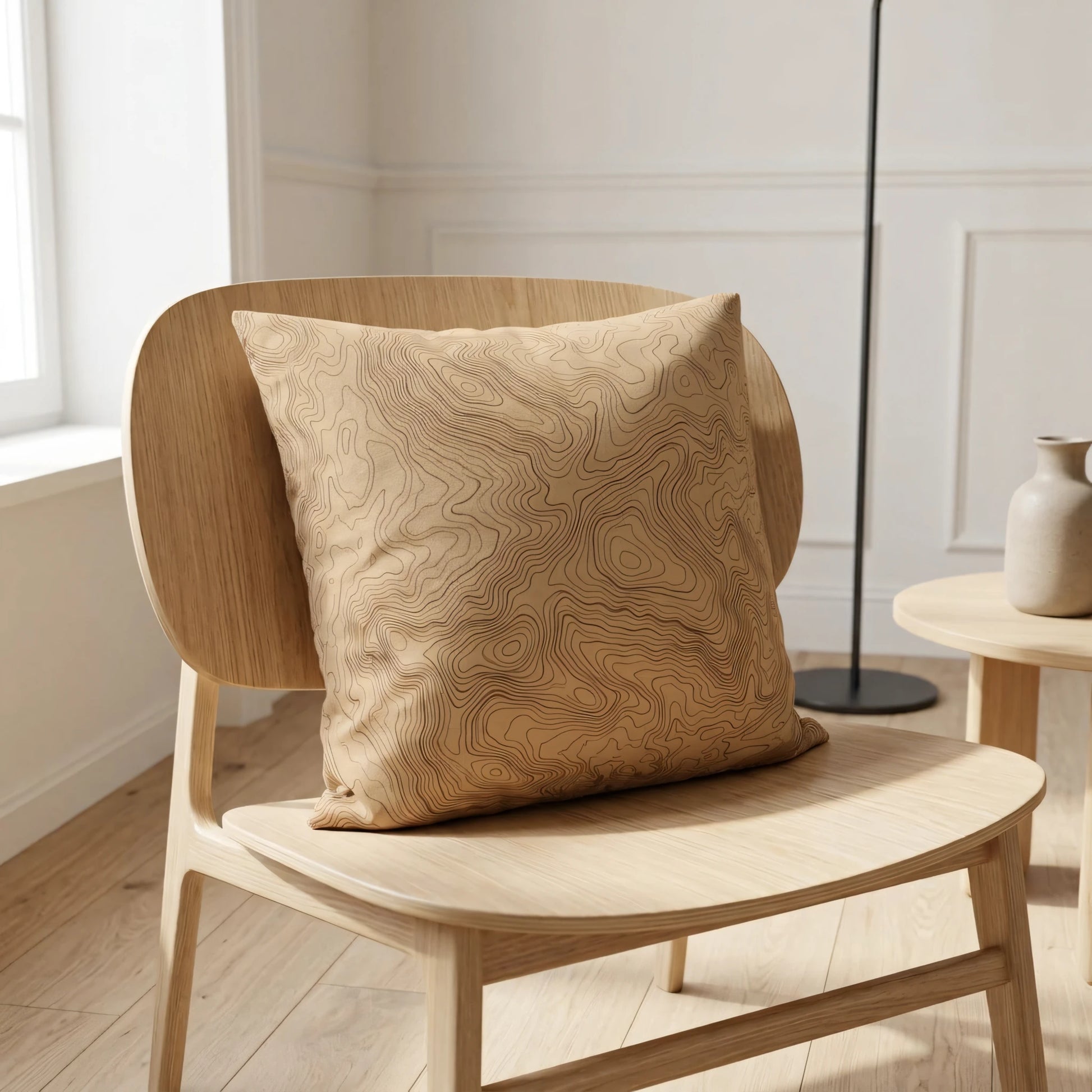 Topographic map pattern pillow in tan on a light oak chair within a minimalist interior featuring soft natural lighting.